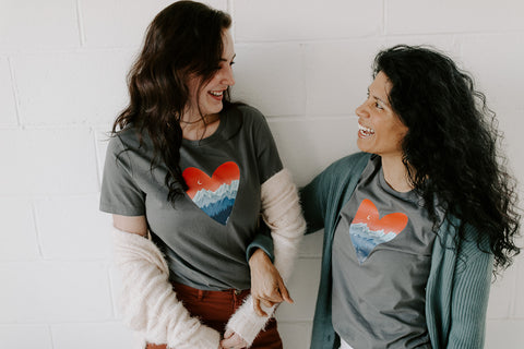 Two women wearing grey t-shirts with a heart with mountains 