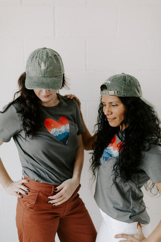 Two women wearing grey t-shirts with hearts and mountains and wearing embroidered hats with pine trees