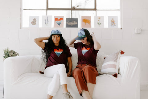 Two women sitting on a couch with art prints behind them wearing t-shirts and trucker hats