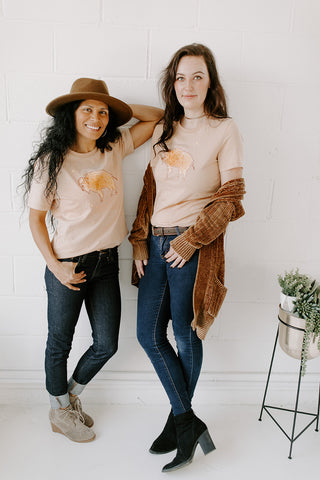 Two women wearing beige t-shirts with bison/buffalo on them sweater wool hat jeans and t-shirt casual