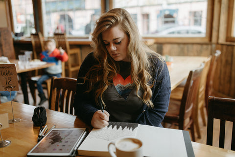 Shelby Campbell sitting in Leavenworth coffee shop wearing a grey graphic t-shirt with a heart and mountain scene illustration. PNW Style. Pacific Northwest. Drawing. Sipping Coffee. Cafe.  Leavenworth, Washington. Mountains. Mountain Style.