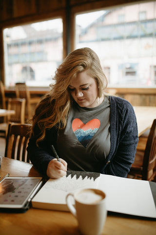 Shelby Campbell sitting in Leavenworth coffee shop wearing a grey graphic t-shirt with a heart and mountain scene illustration. PNW Style. Pacific Northwest. Drawing. Sipping Coffee. Cafe.  Leavenworth, Washington. Mountains. Mountain Style.