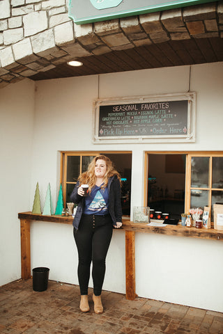 Women wearing a blue graphic t-shirt with a triangle shaped mountain illustration on the front. Black denim jeans. High Waisted. Booties. Coffee. Outdoor Coffee Shop in Leavenworth, Washington. PNW Style. Pacific Northwest.