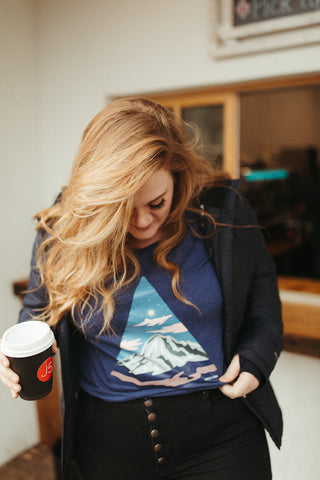 Women wearing a blue graphic t-shirt with a triangle shaped mountain illustration on the front. Black denim jeans. High Waisted. Booties. Coffee. Outdoor Coffee Shop in Leavenworth, Washington. PNW Style. Pacific Northwest.