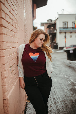 Mountain Love Graphic T-shirt. Tee. Mountain and Heart Illustration. Woman wearing white sweater, black pants, and brown booties in a snowy forested area in Leavenworth, Washington. PNW Style. Pacific Northwest. Casual Style. Mountain Style. Mountains.