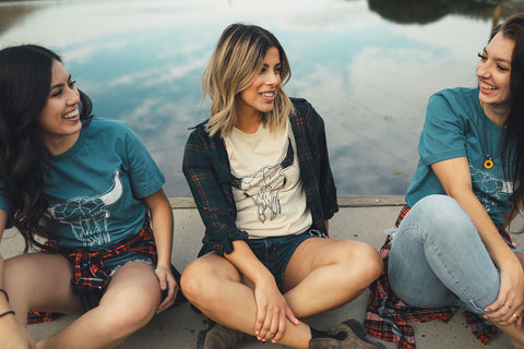 Group of women sitting in front of a small lake/pond. Wearing graphic t-shirts with a steer skull design on the front. PNW style. Wenatchee, WA. Pacific Northwest. Graphic Tee.