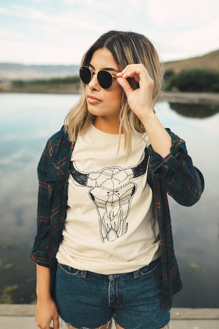 Woman standing in front of small lake/pond in Wenatchee, WA. Wearing a graphic t-shirt with a steer skull design on the front. Jeans Shorts. Round Sunglasses. Summer Time.PNW Style. Pacific Northwest.