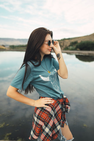 Woman standing in front of small lake/pond in Wenatchee, WA. Wearing a graphic t-shirt with a steer skull design on the front. Jeans. Flannel Shirt. Round Sunglasses. Summer Time.PNW Style. Pacific Northwest.