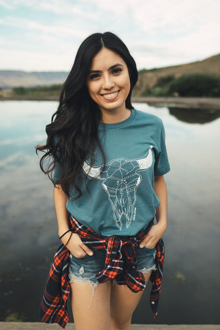 Woman standing in front of small lake/pond in Wenatchee, WA. Wearing a graphic t-shirt with a steer skull design on the front. Jeans. Flannel Shirt.  Summer Time.PNW Style. Pacific Northwest.