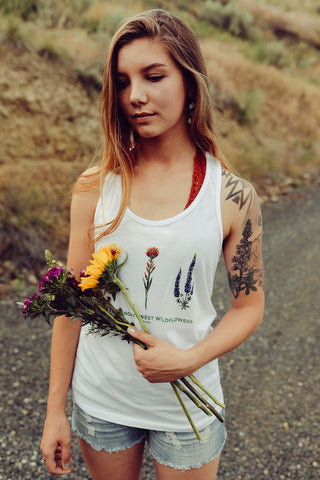 Woman standing in front of a dirt road wearing a white tank top featuring an illustration of Pacific Northwest Wildflowers. She is holding a bouquet of flowers. Jeans shorts. Summer time. Flowers. Floral. Wildflower. Wenatchee, Washington. PNW. Pacific Northwest Style.