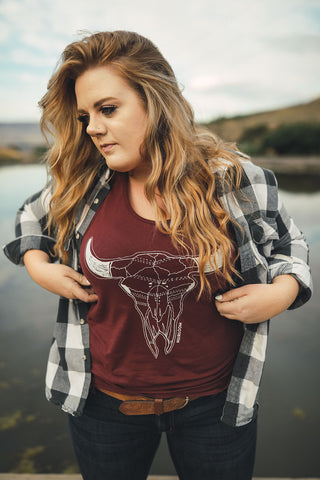 Woman standing in front of small lake/pond in Wenatchee, Washington. She is wearing a maroon colored tank top with a steer skull design on the front and jeans with a brown belt, and a flannel shirt. PNW. Pacific Northwest Style.