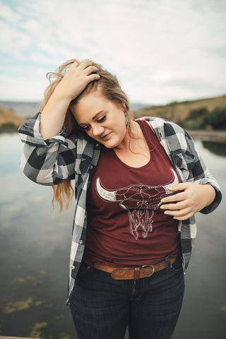 Woman standing in front of small lake/pond in Wenatchee, Washington. She is wearing a maroon colored tank top with a steer skull design on the front and jeans with a brown belt, and a flannel shirt. PNW. Pacific Northwest Style.