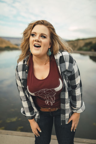 Woman standing in front of small lake/pond in Wenatchee, Washington. She is wearing a maroon colored tank top with a steer skull design on the front and jeans with a brown belt, and a flannel shirt. PNW. Pacific Northwest Style.
