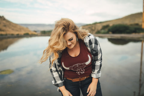 Woman standing in front of small lake/pond in Wenatchee, Washington. She is wearing a maroon colored tank top with a steer skull design on the front and jeans with a brown belt, and a flannel shirt. PNW. Pacific Northwest Style.