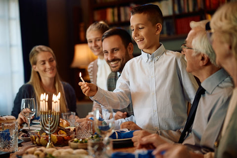 Happy Jewish extended family celebrating Hanukkah gathering at dining table. Focus is on kid lighting candles in menorah.