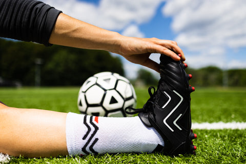 player sits on football pitch with outstretched leg and reaches for toes to stretch while wearing Ida Sports football boots