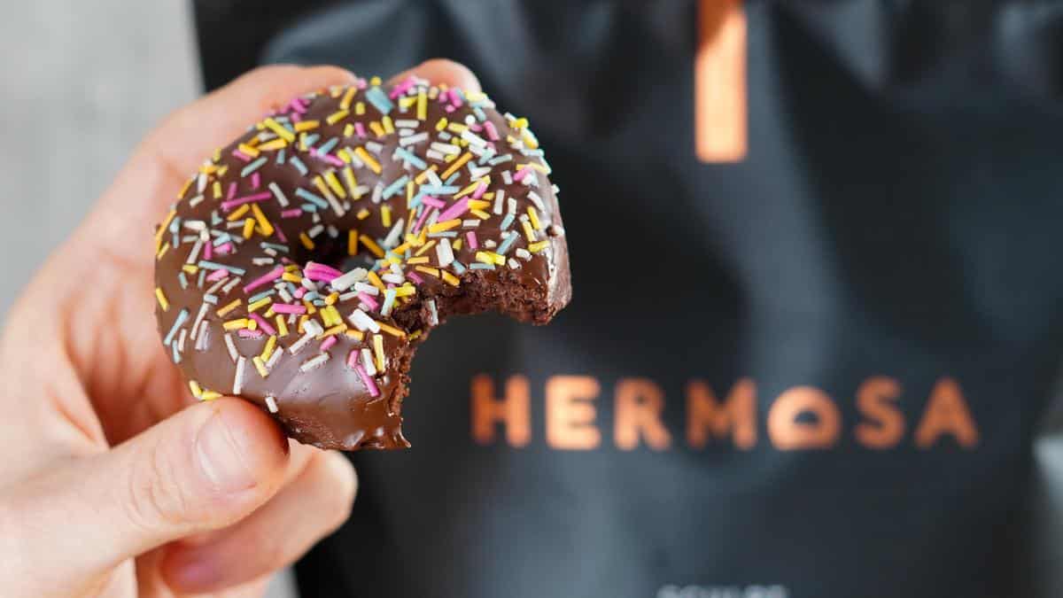 A chocolate protein doughnut being held in front of a bag of HERMOSA protein powder.