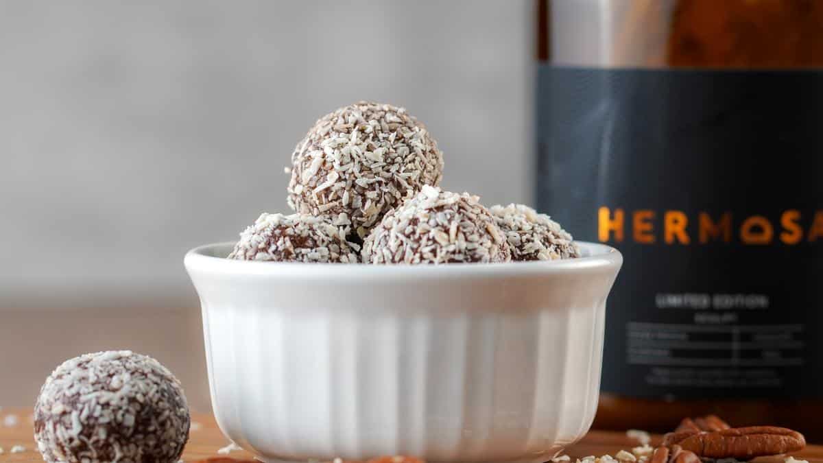 Protein energy balls in a white cup with a HERMOSA protein powder glass jar in the background.