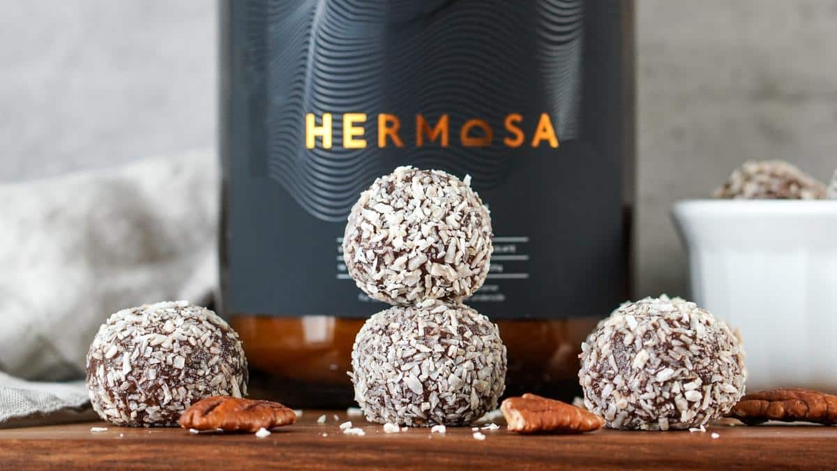 Four protein energy balls in front of a glass jar of HERMOSA protein powder.