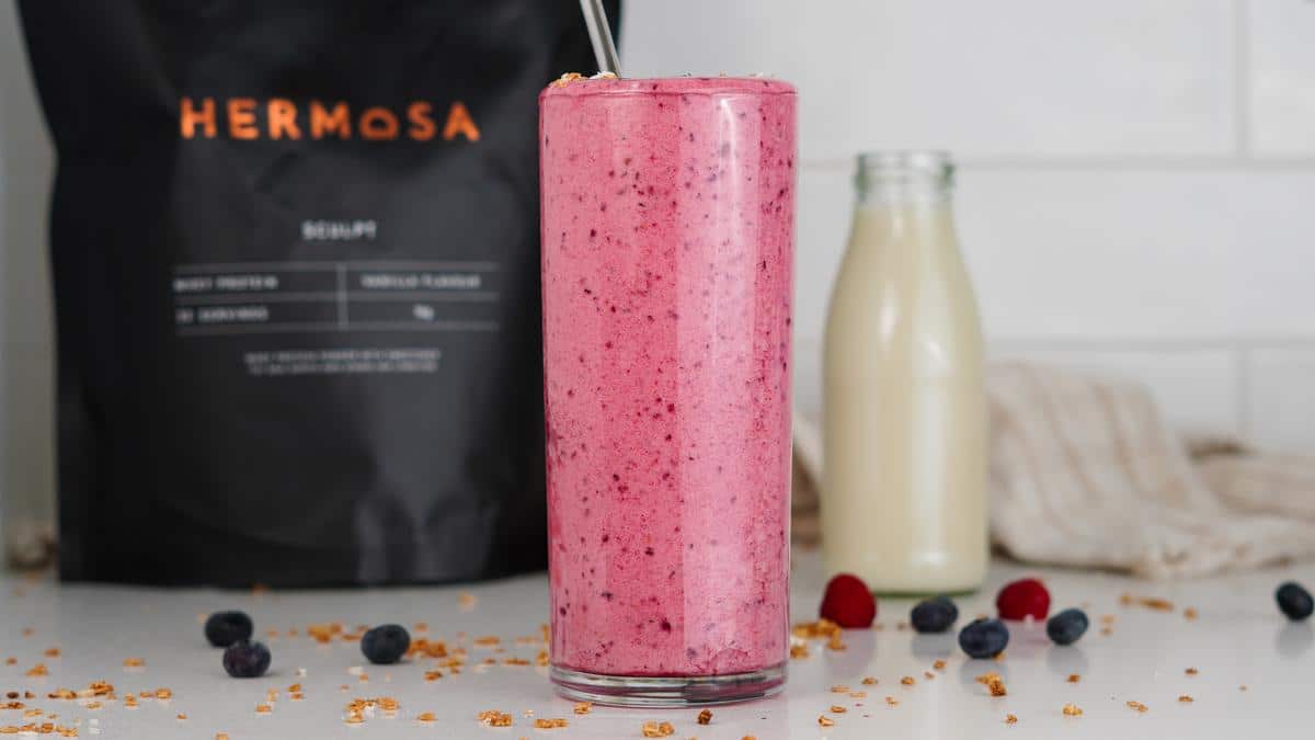 A berry protein smoothie next to a glass milk bottle and with. bag of HERMOSA protein powder in the background.
