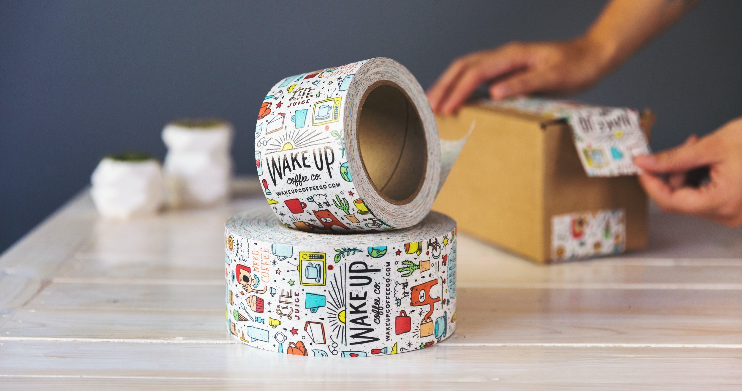 Branded packing tape is stacked on a table with a person taping a box behind it.