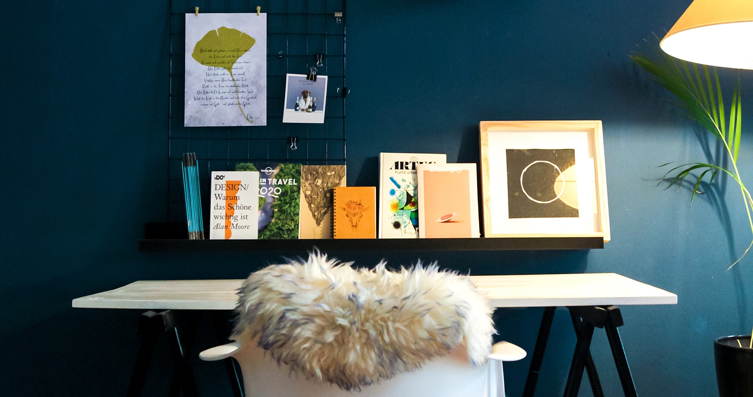 A small workspace against a blue wall, lit by a floor lamp and featuring a number of art prints