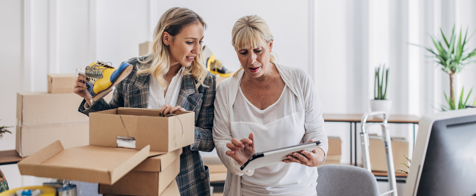 Two women preparing orders to ship to customers. One of them is putting a shoe inside a box and they are both looking at a tablet at the same time.