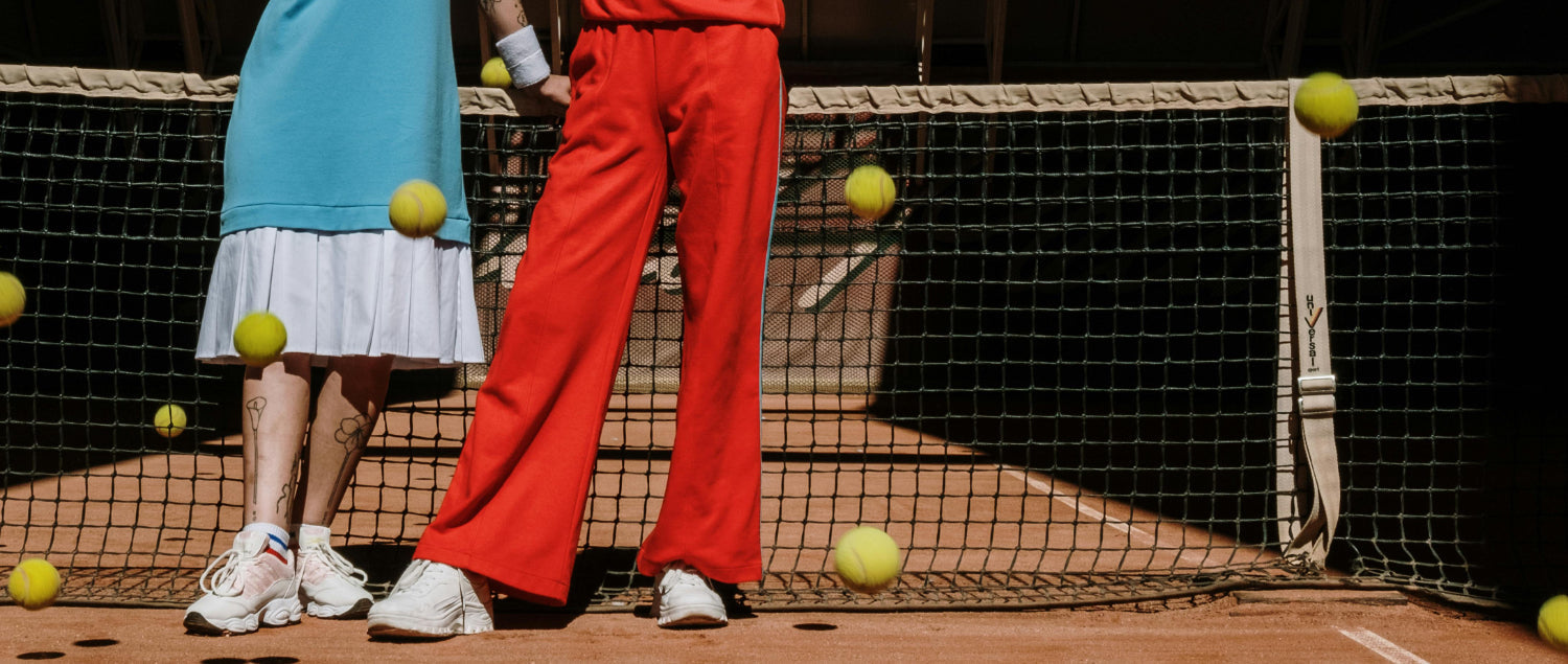 Two people stand in front of a tennis net and tennis balls fly around