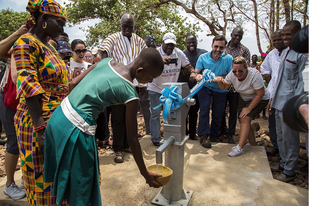 A group of individuals surrounding a well with a bright blue bow on top as a teenager pours water from the well. 