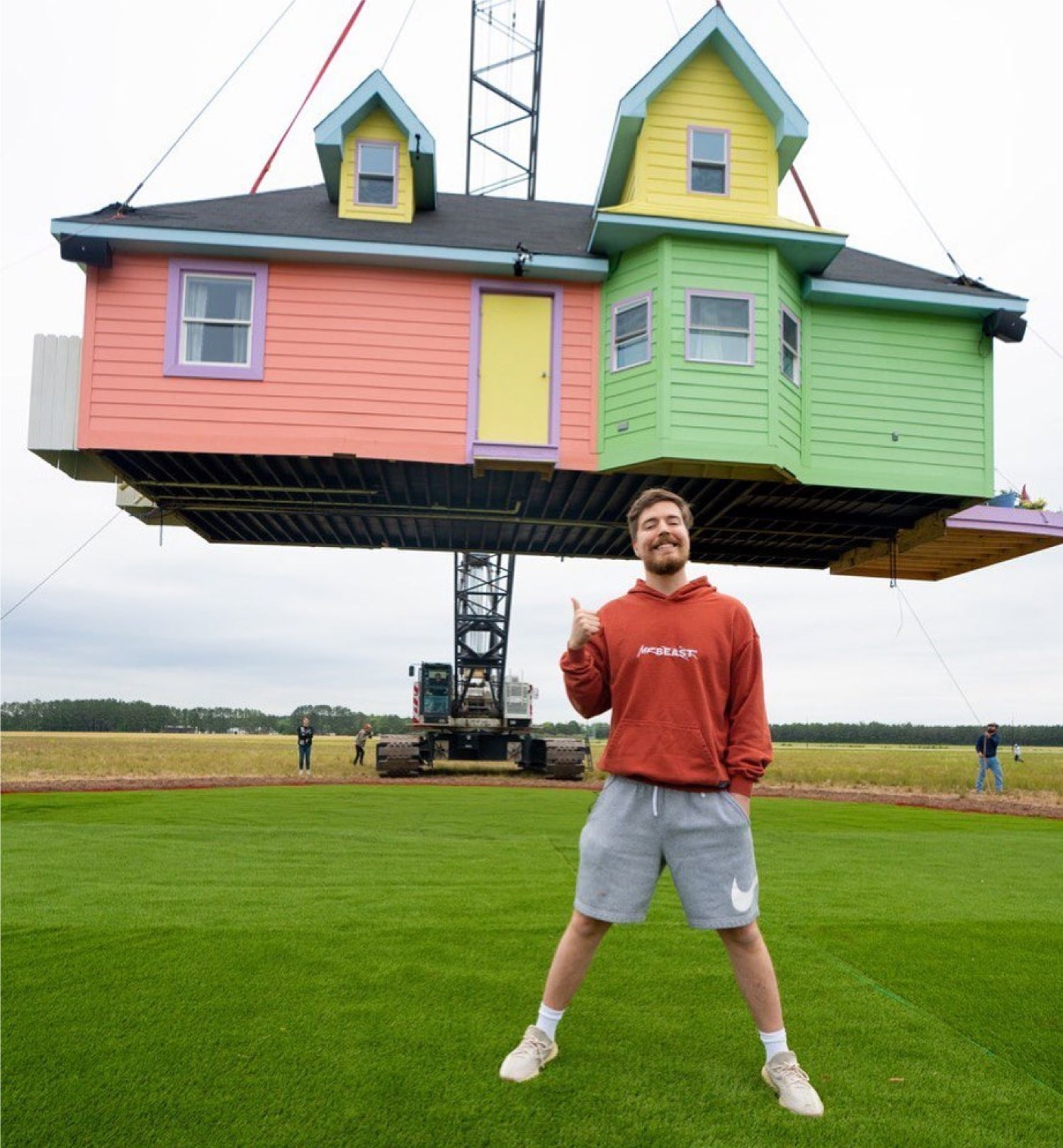 MrBeast stands in an open field wearing a red hoodie and gray shorts. He is smiling and giving the camera the thumbs-up sign. Above him, a crane suspends a whimsical multi-colored house.