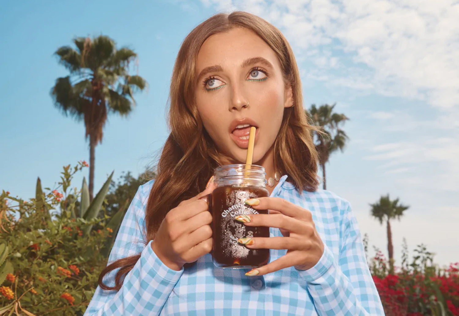 A close-up photo of Emma Chamberlain standing outside while looking up into the sky and sipping on Chamberlain Coffee’s Mason Jar Cold Brew.