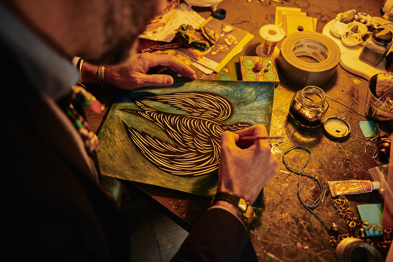 Detail of a man’s hands painting a bird design onto a canvas. The table is covered with art supplies.