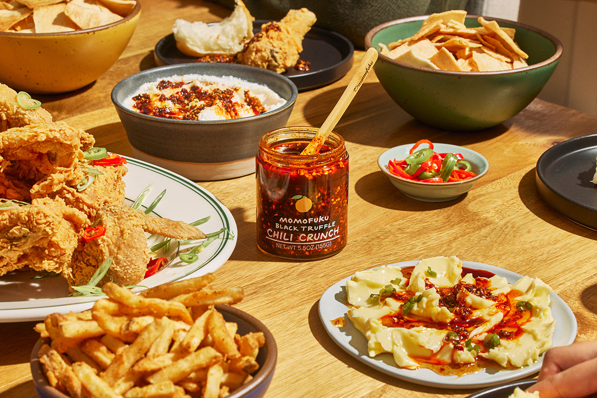 A jar of Momofuku black truffle chili crunch surrounded by dishes of fries, chicken wings, chips, dips and eggs