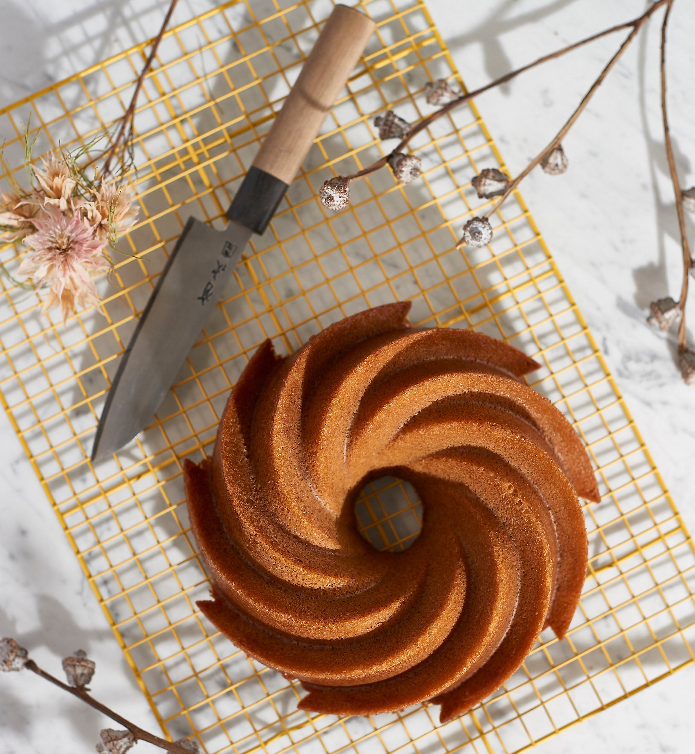 a wild rye cake sitting on a yellow baking rack with a knife and flowers laying around it.