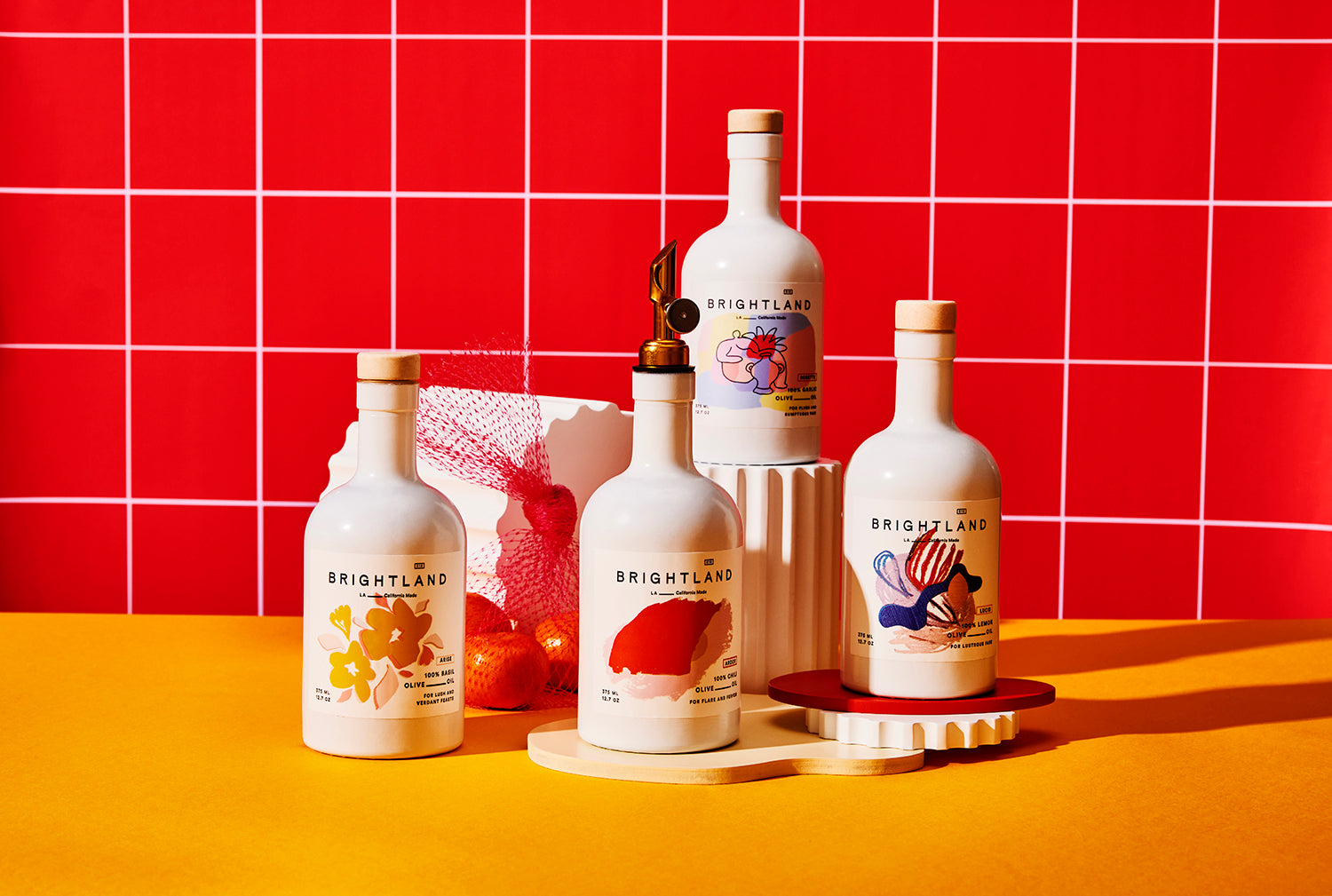 Four Brightland white olive oil bottles with bright graphics on a counter against red tile.