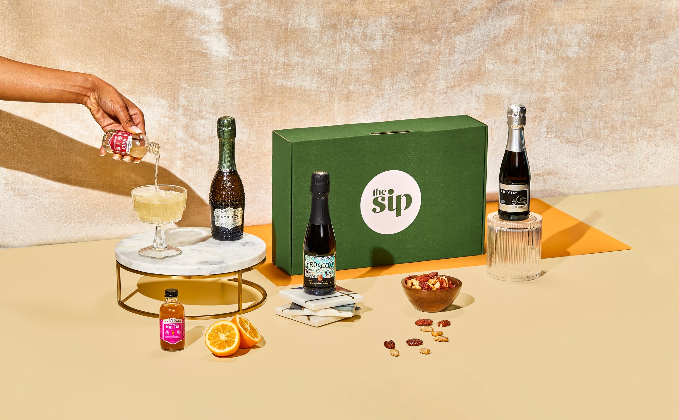 A hand pours the contents of the simple mule into a class, with The Sip’s curated box on display and bottles and fruits on display. 