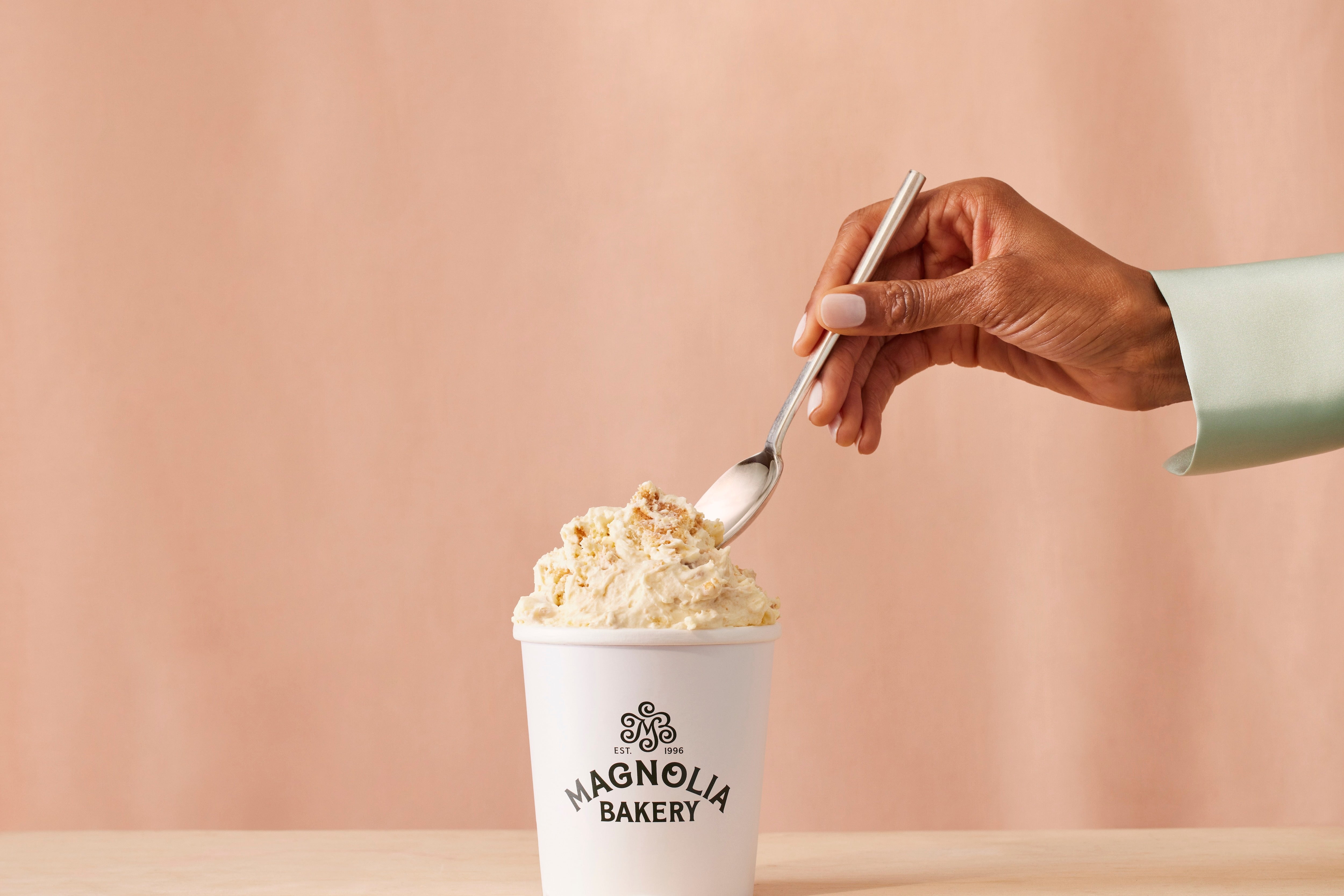 a hand reaches toward a magnolia bakery banana pudding with a silver spoon in hand.