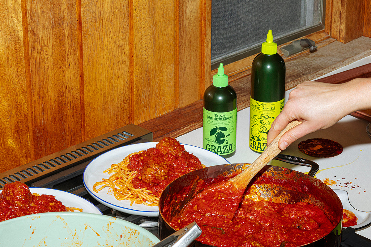 Two bottles of Graza olive oil next to a person making spaghetti in a pan