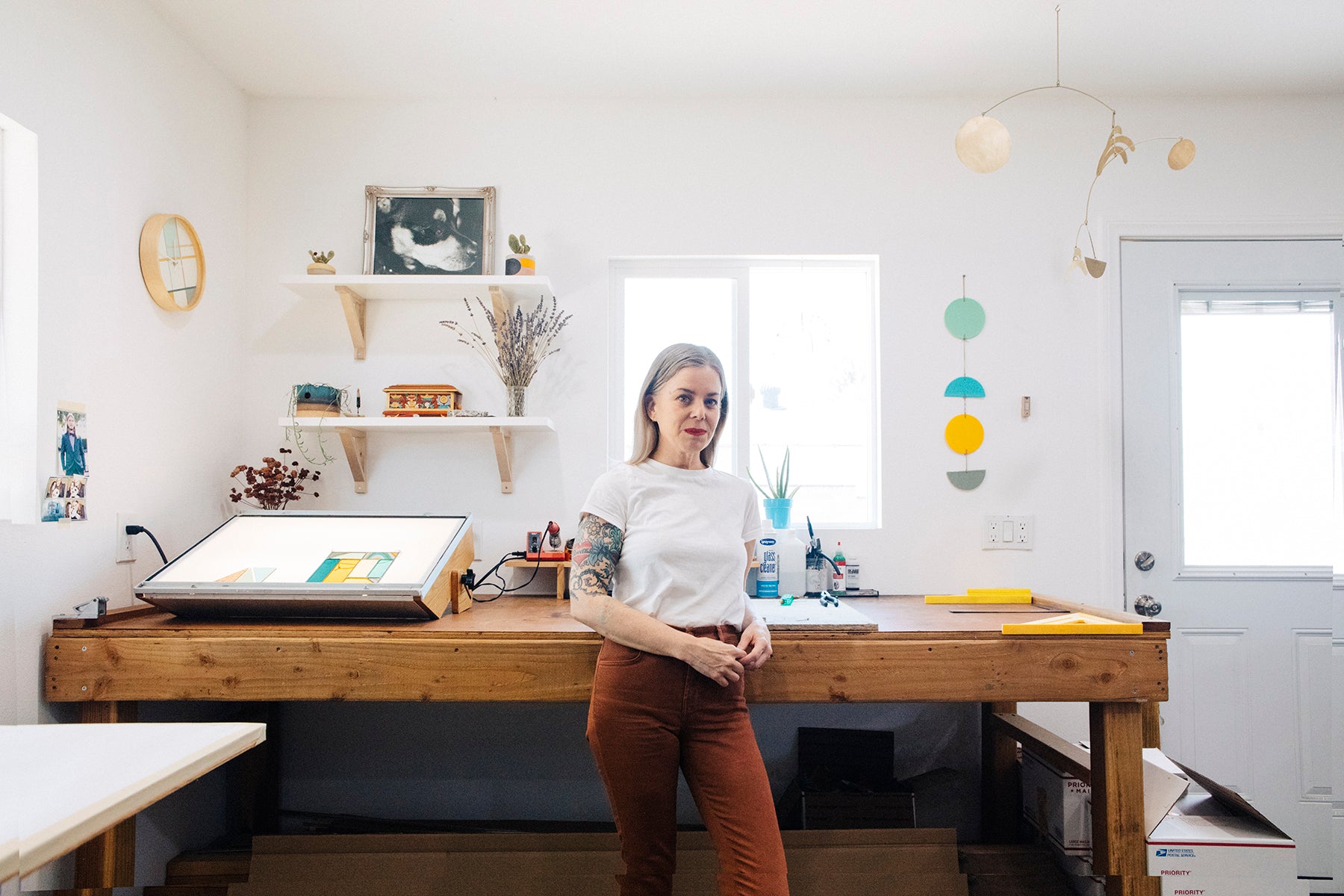 Portrait of artist Debbie Bean today, pictured in her current studio. She is wearing a white t shirt and brown jeans and she is resting against a large wood surface which has a light table on it. Samples of her stained glass and other personal items are hanging on the wall behind her. 