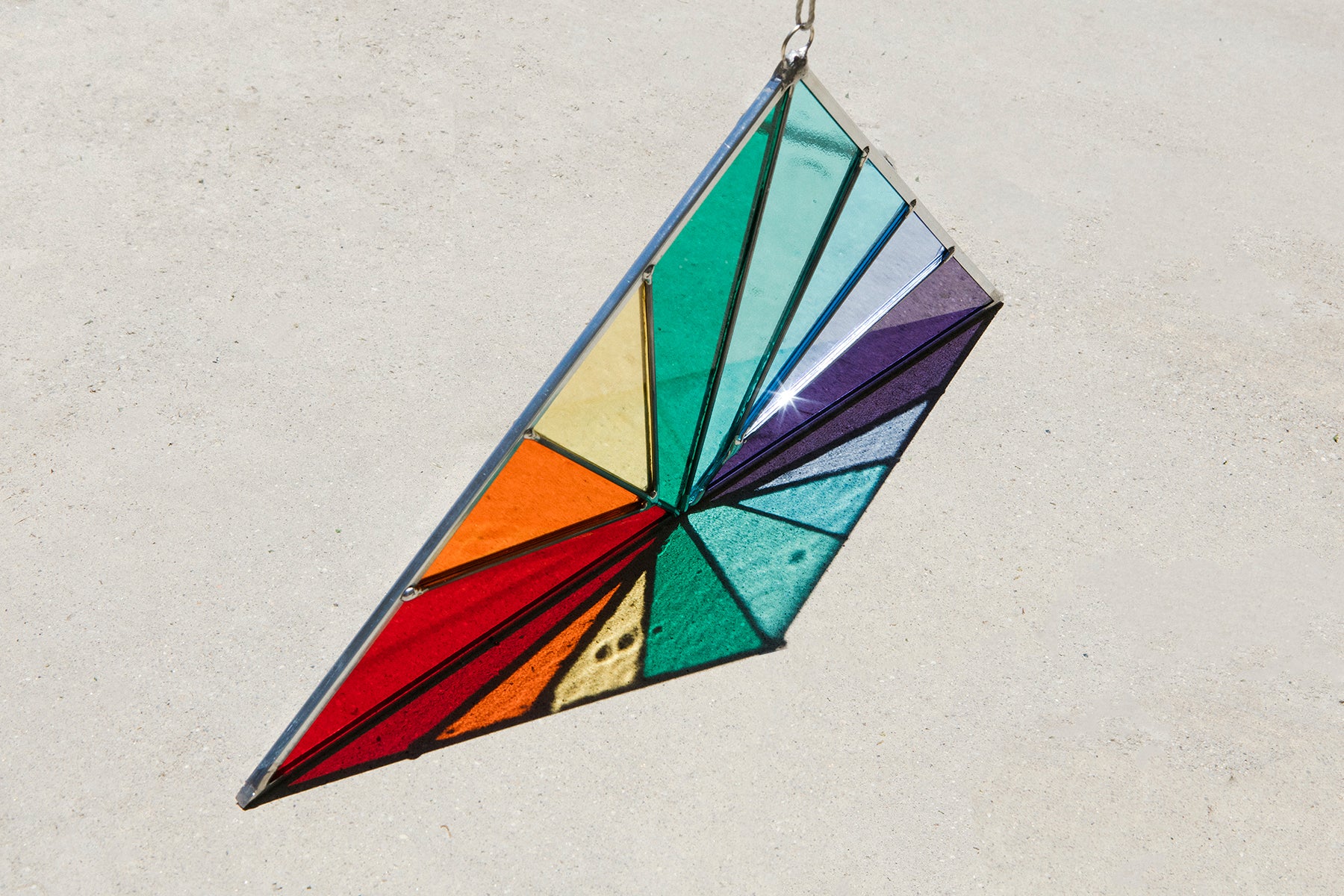 Photograph of a larger, multi coloured, geometric stained glass piece made by Debbie Bean, hanging outside with the sunlight shining through it. 