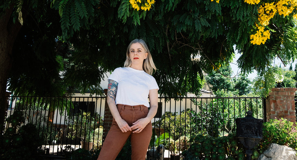 Portrait of the artist Debbie Bean standing under a tree with yellow flowers wearing a white t shirt and brown jeans with red lipstick on.