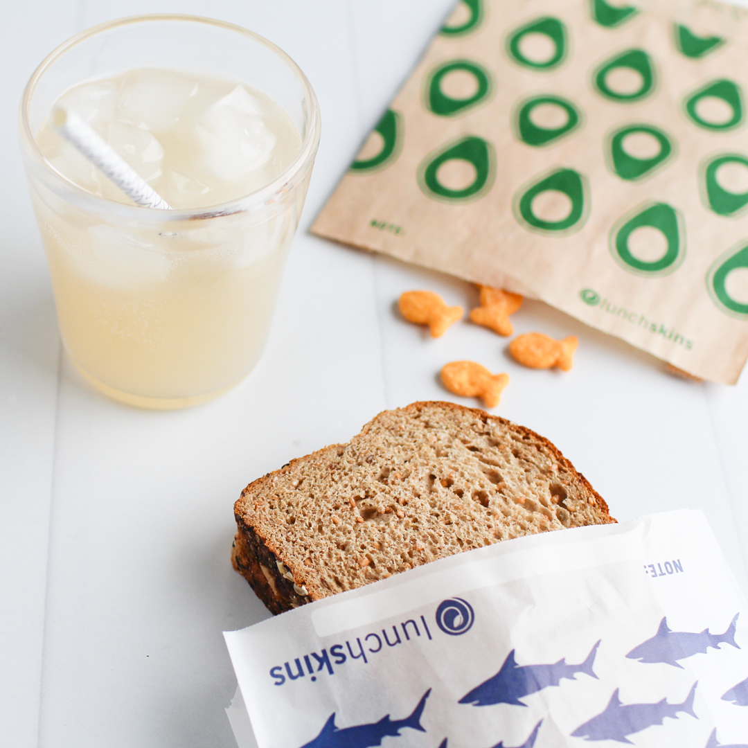 Two Lunchskins compostable, unbleached snack bags with resealable stickers holding a sandwich and goldfish next to a Lunchskins biodegradable straw. 