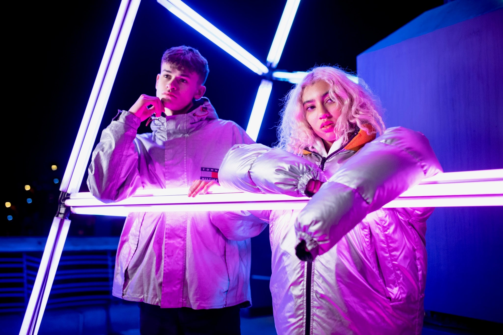 A male and female model wears puffy winter vintage jackets surrounded by neon lights. 