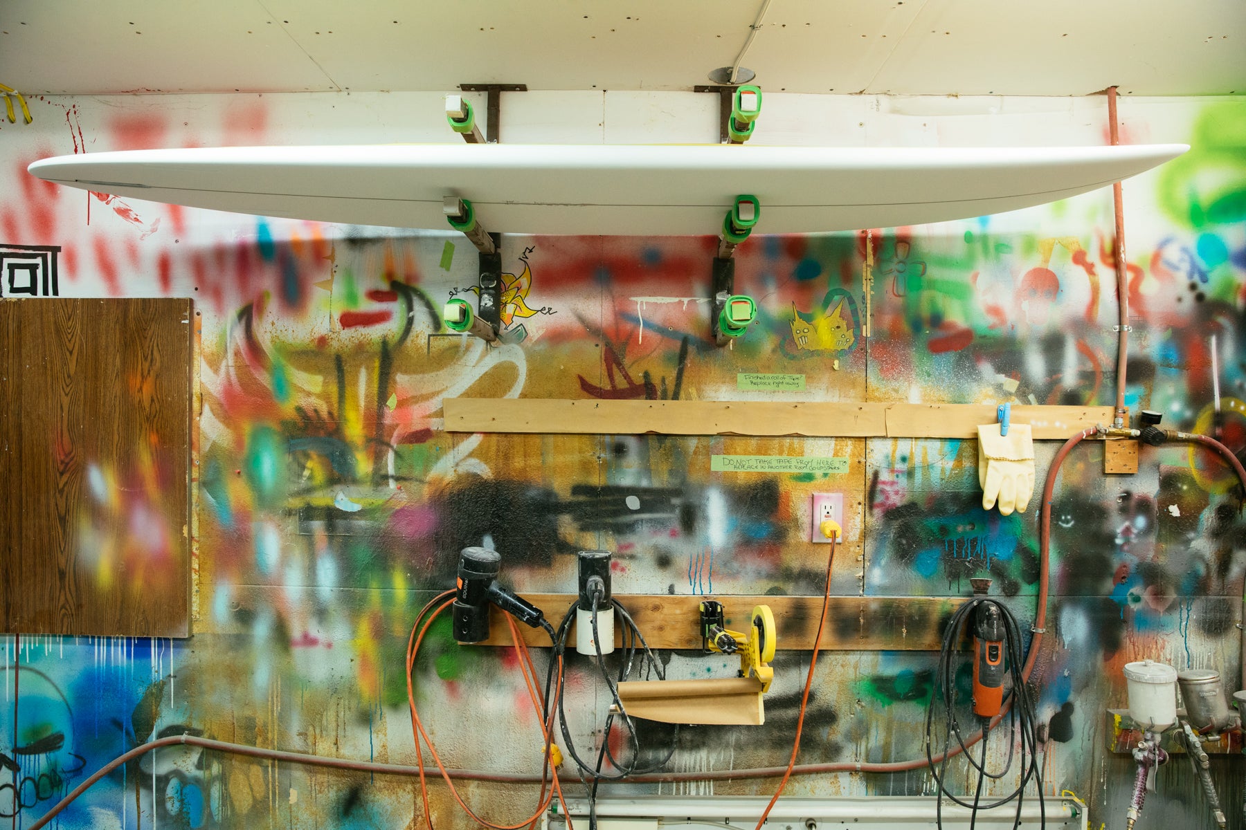A blank surfboard hangs on a top shelving unit against a colourful spray painted backdrop in the Aftanas workshop. 