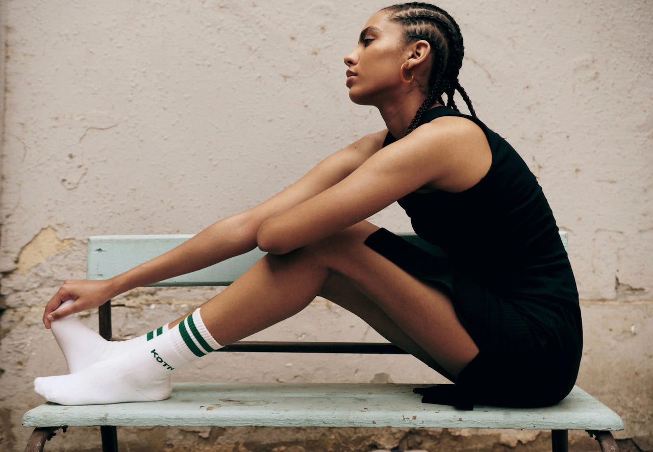 A model in a black dress and white socks by KOTN sitting on a pale green bench.