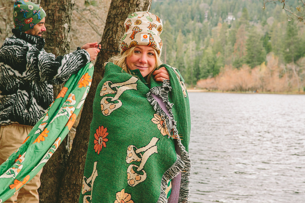 Woman wrapped in a blanket and wearing a hat standing next to a body of water