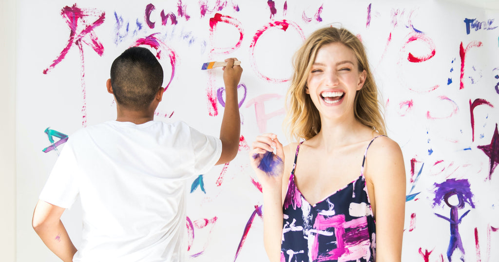 Woman in a printed dress laughs in the foreground while a man paints a wall behind her