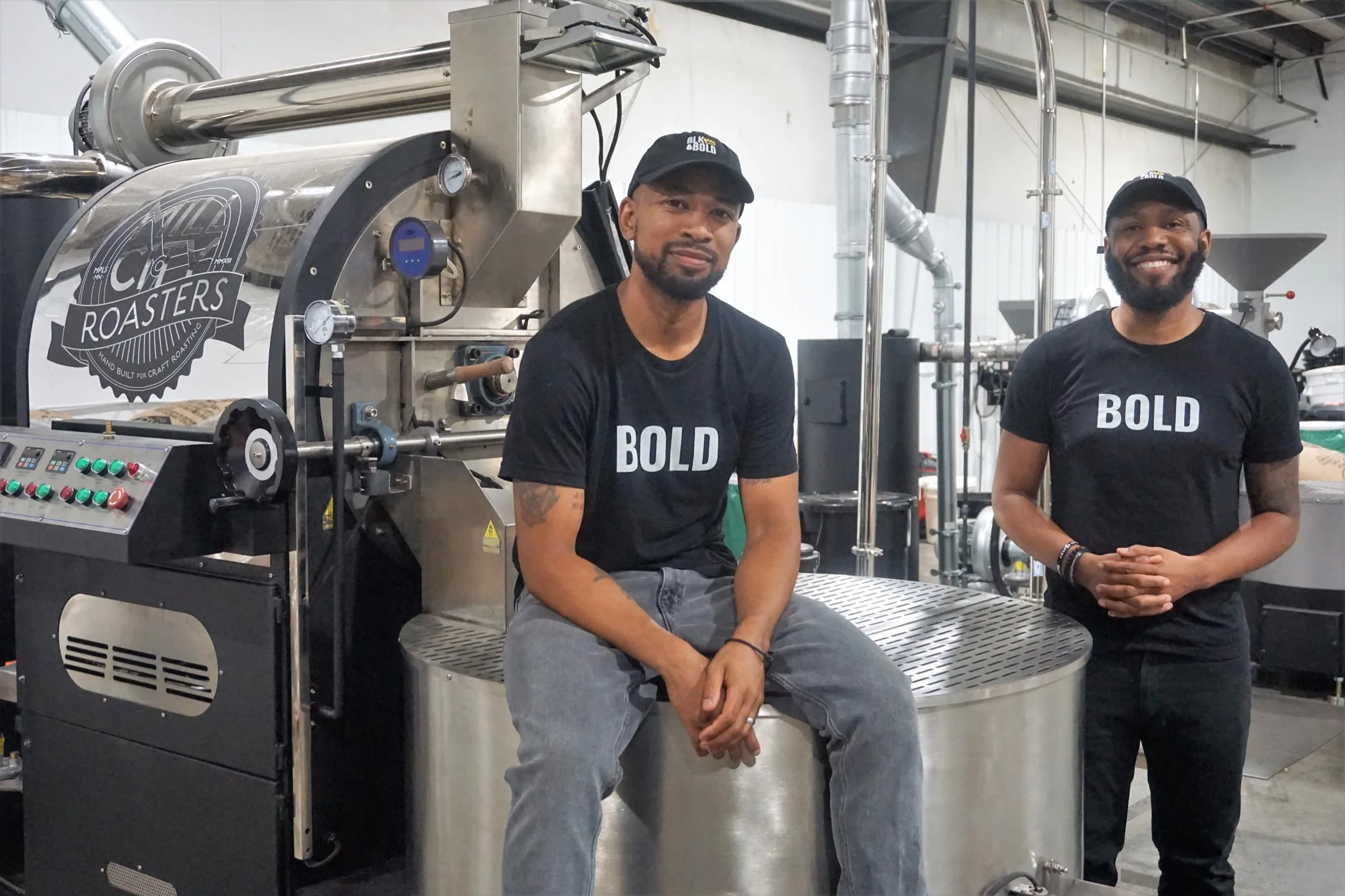 Two men in t-shirts, sitting on and leaning on a coffee roasting machine smiling.