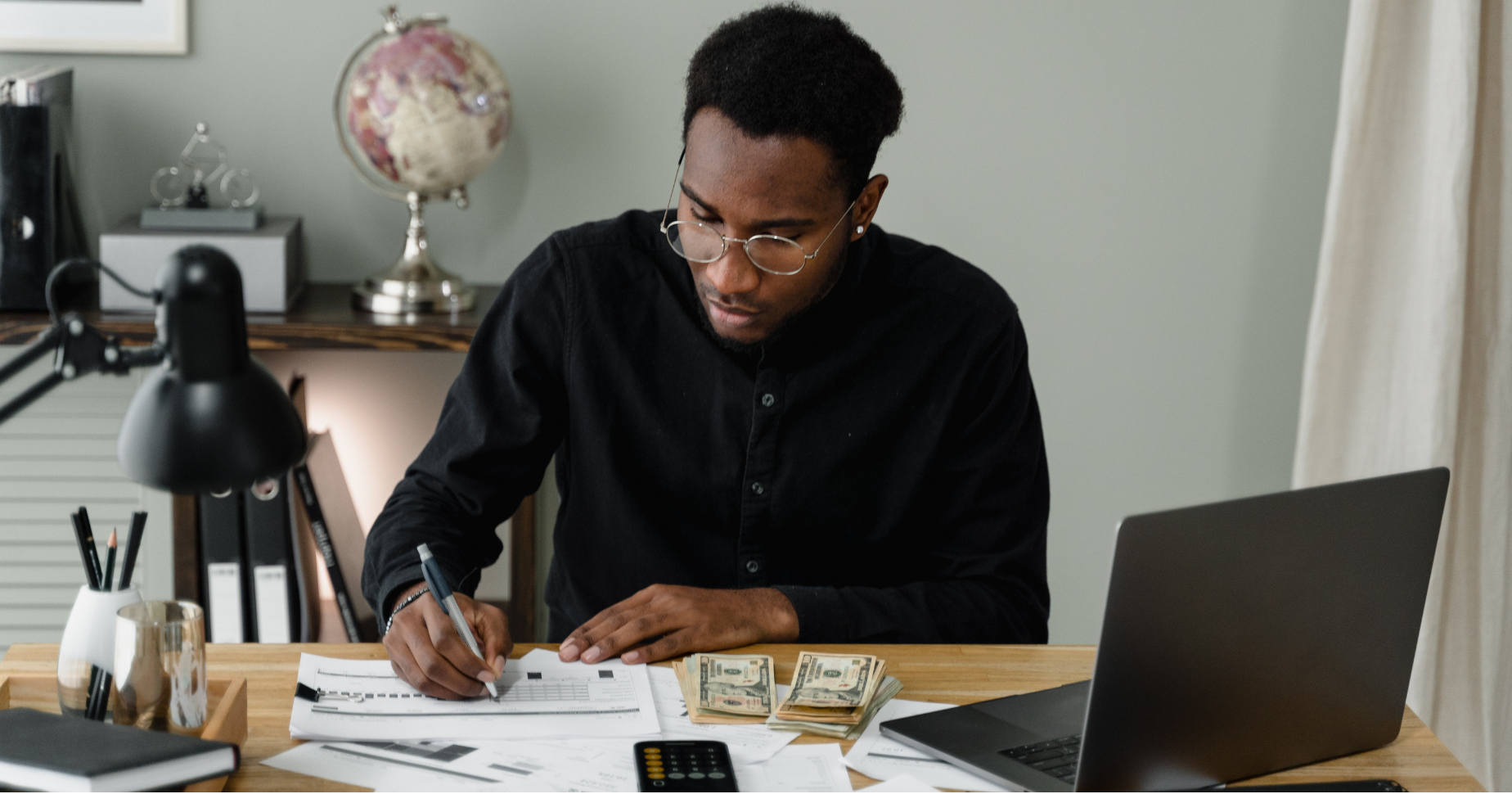 A man works at a desk next to stacks of cash and a laptop, he makes notes on a notebook