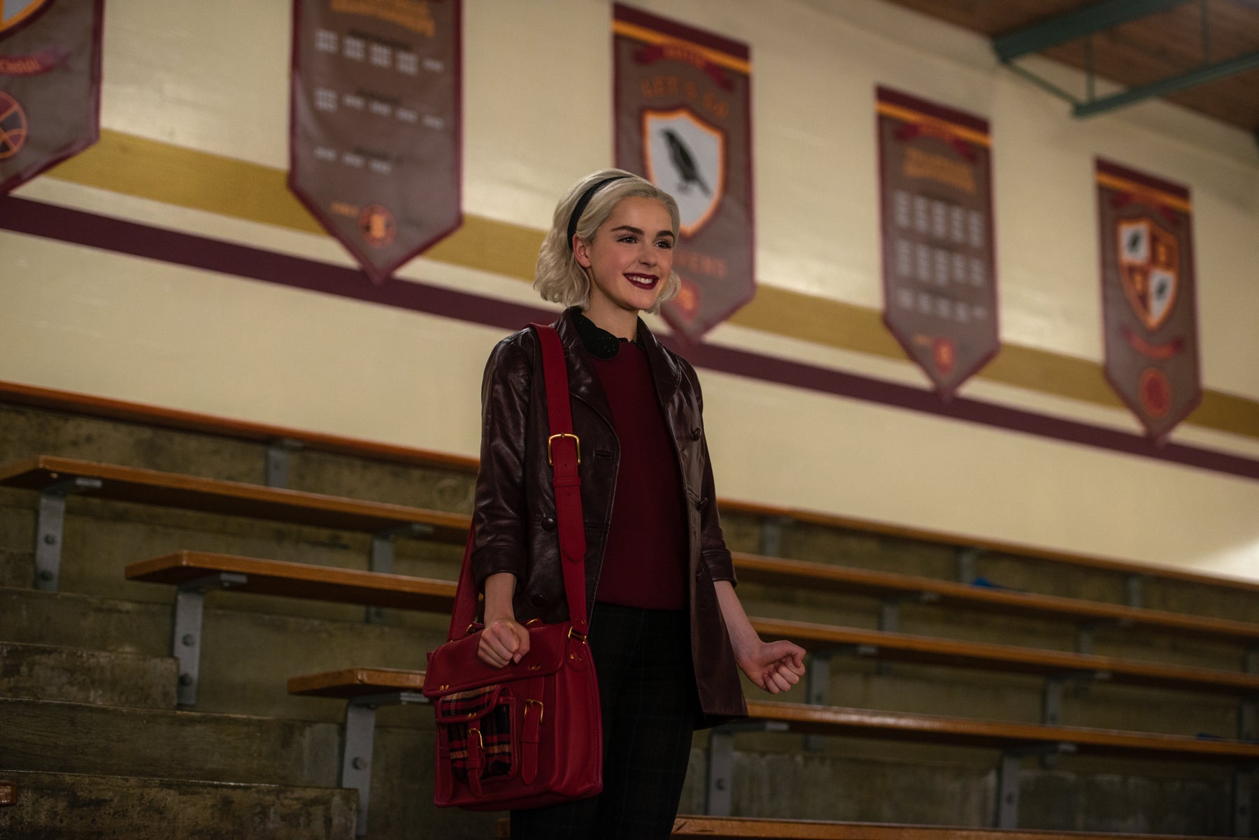 Sabrina Spellman stands smiling in the bleachers of the Greendale High gym in a scene from Chilling Adventures of Sabrina.